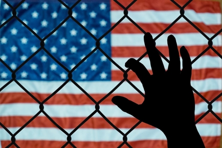 A silhouette of a hand grasps a fence with a U.S. flag blurred in background.