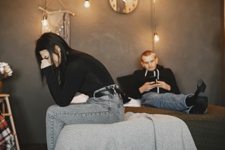 A woman sits on the edge of a bed with her head in her hands, appearing distressed, while a man lies behind her looking at his phone in a dimly lit bedroom.