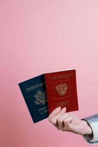 A hand holds two passports, one from the United States and one from the Russian Federation, against a pink background.